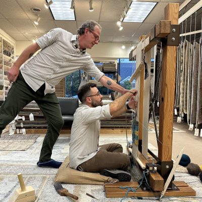 Weaving is a tactile craft better understood through the hands. As such, Michael Christie, The Ruggist, welcomes others to sit, experience, and contemplate 'The Nature of Weaving' during his presentation of the same name. Here, Fouad Mahfuz of PRG Rugs in Nashua, New Hampshire tries his hand at weaving under the supervision of The Ruggist. | Photograph courtesy of PRG Rugs, 21 September 2023
